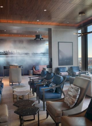 The image shows a modern lounge with seating, large windows, and a cityscape mural on the wall. It has a view of water and mountains.