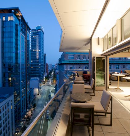 A modern balcony with seating overlooks a cityscape of tall buildings and illuminated streets at dusk.