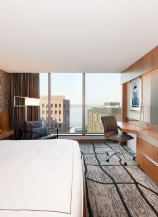 A modern hotel room with a large bed, wall-mounted TV, desk, chair, and window view of buildings. The decor is sleek and stylish.