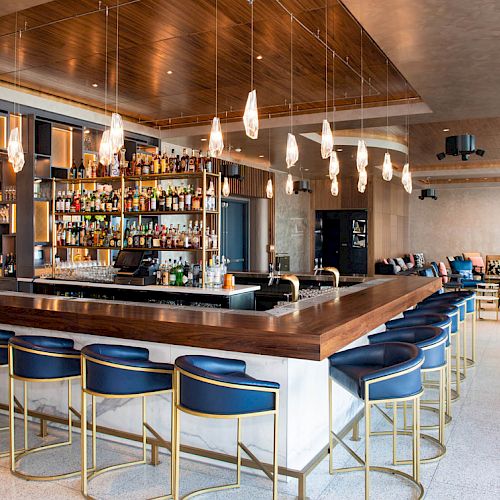 The image shows a modern bar with a wooden counter, blue bar stools, pendant lights, and a stocked liquor shelf in a chic indoor setting.