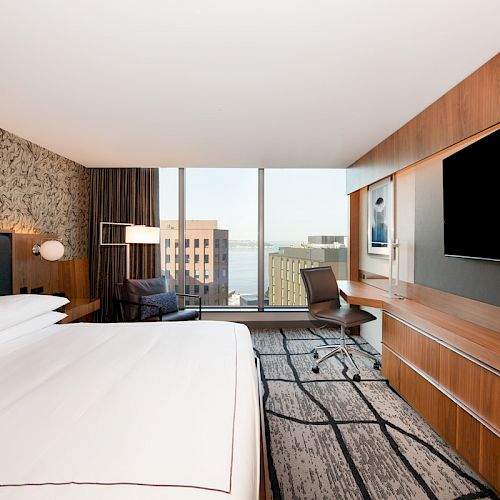 A modern hotel room with a large bed, TV, desk, and window view of buildings and sky, featuring contemporary design elements.