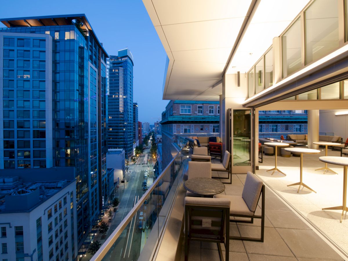 A modern cityscape with tall buildings and a balcony featuring tables and chairs, photographed during the evening.