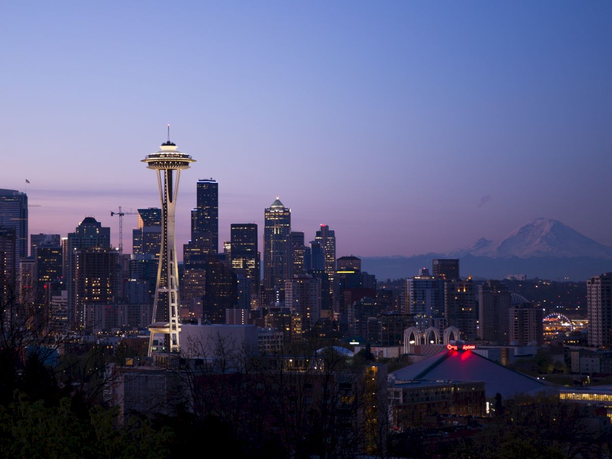 Seattle skyline at dusk, featuring the Space Needle and Mount Rainier in the background, under a soft, dimming sky.