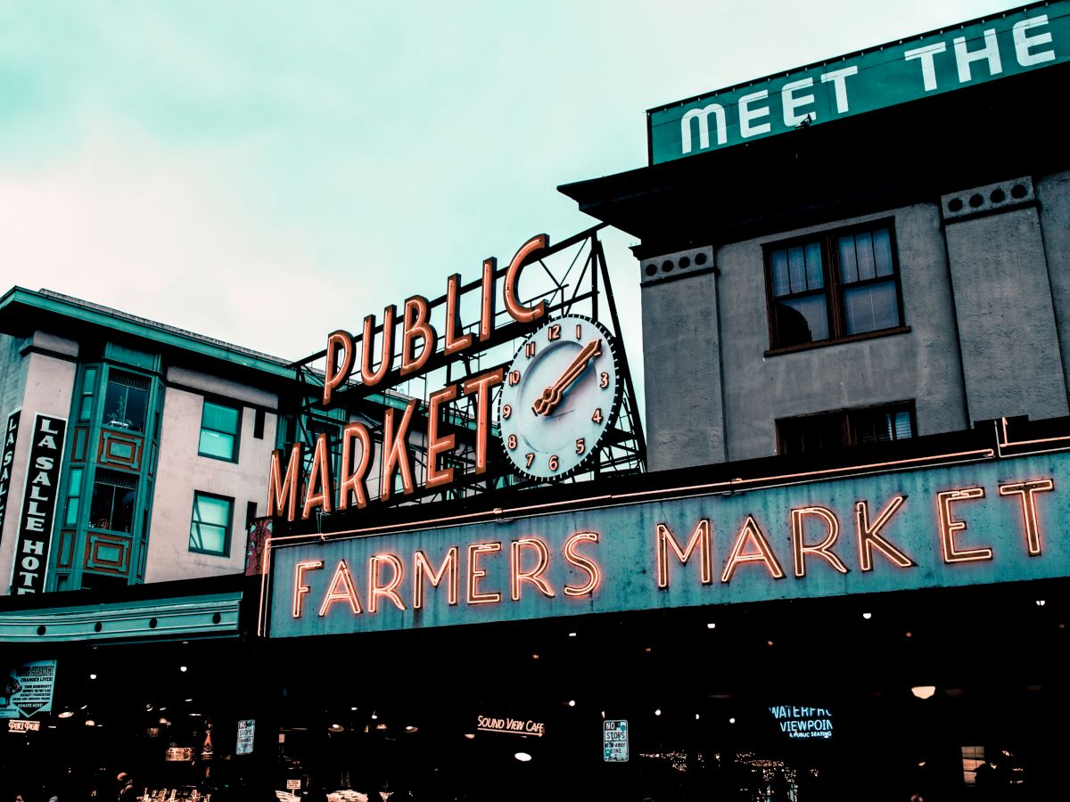 The image shows the iconic neon sign for a public farmers market with a clock, against a city backdrop.