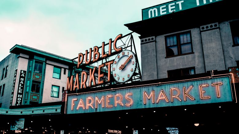 The image shows the iconic neon sign for a public farmers market with a clock, against a city backdrop.