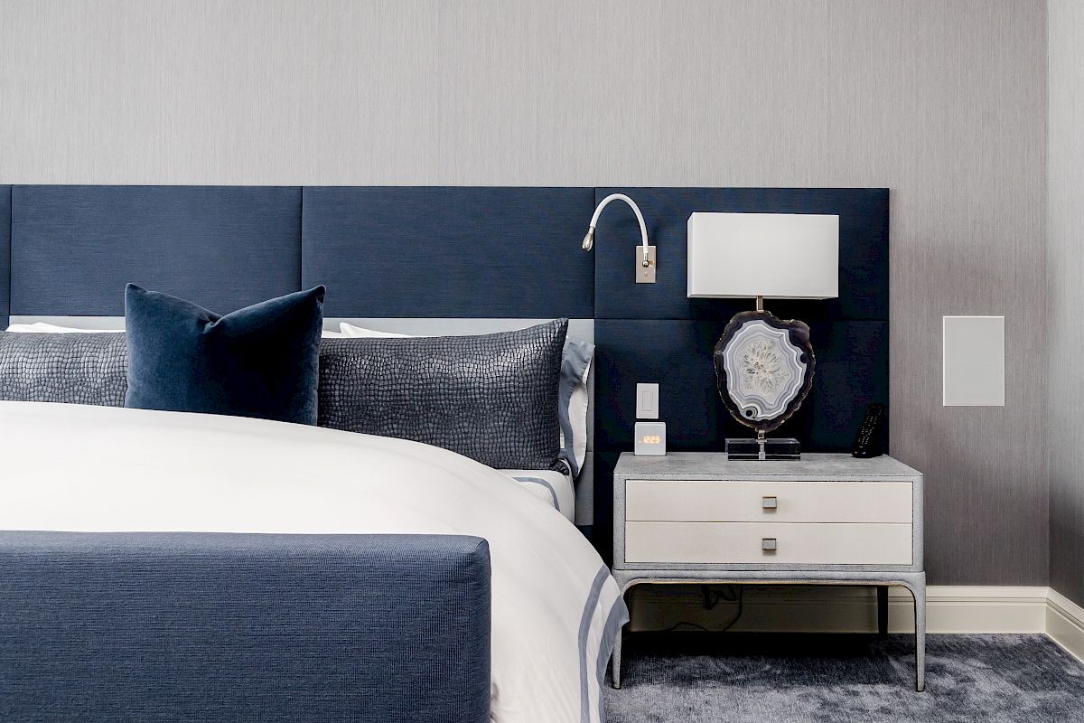 Modern bedroom with a blue and gray theme, featuring a bed, pillows, nightstand, lamp, and decorative accessories in a neat arrangement.