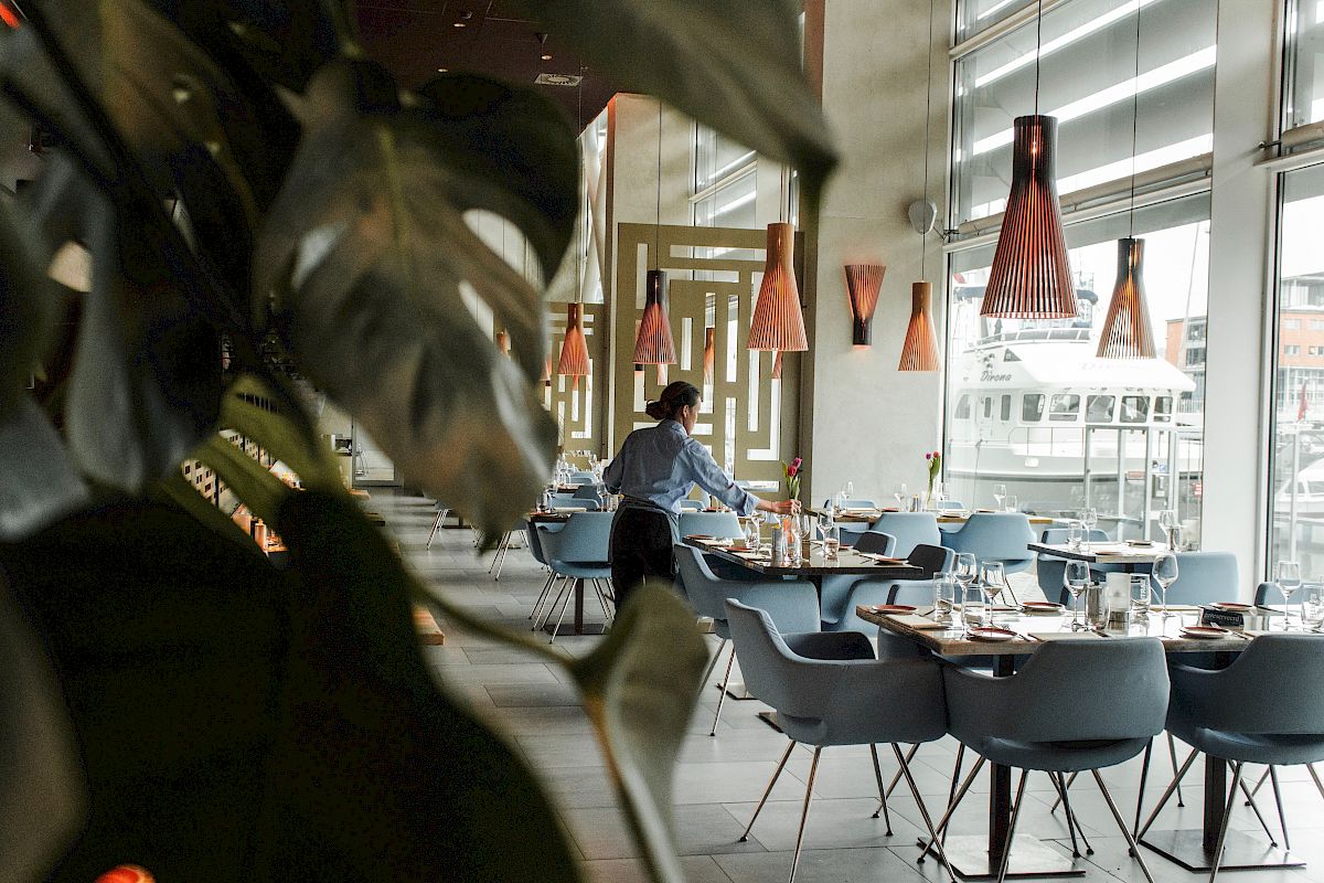 A person is setting tables in a modern restaurant with stylish lamps and large windows. Greenery is partially visible in the foreground.