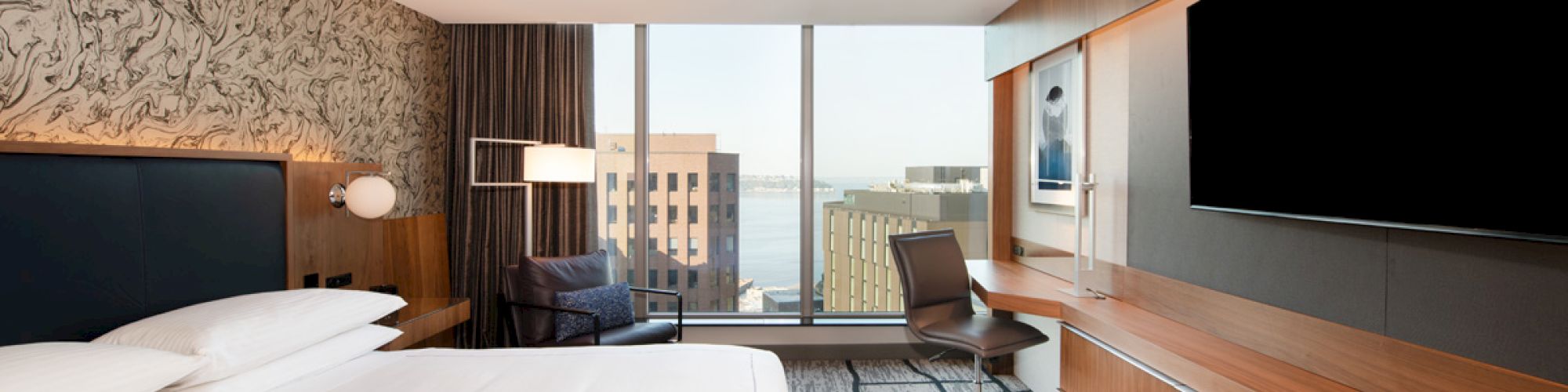 The image shows a modern hotel room with a bed, desk, large TV, and a window offering a city view.